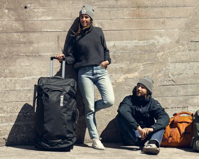 Two people lean against a concrete wall holding items of checked luggage.