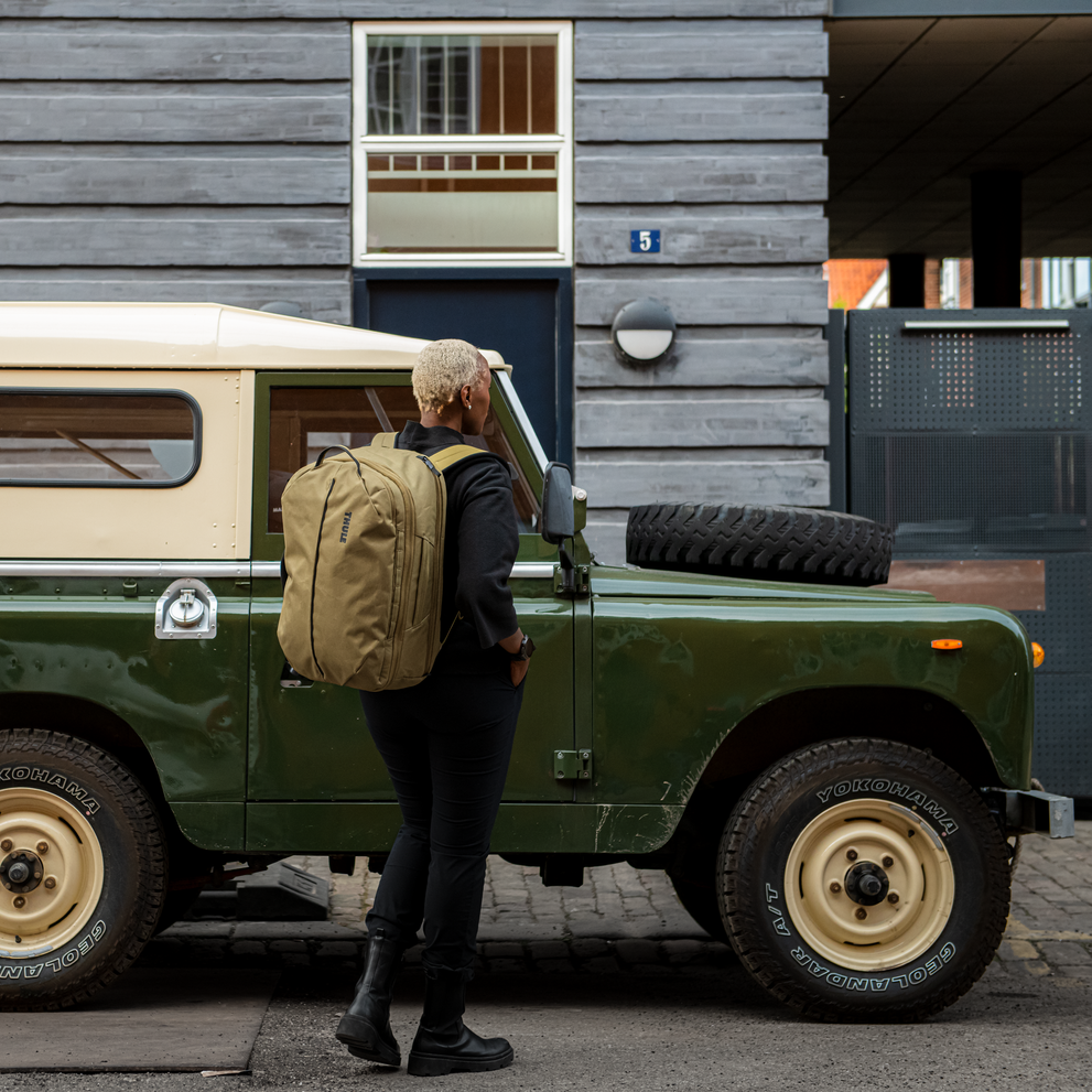 A woman stands next to a green car carrying a tan Thule Aion travel backpack.