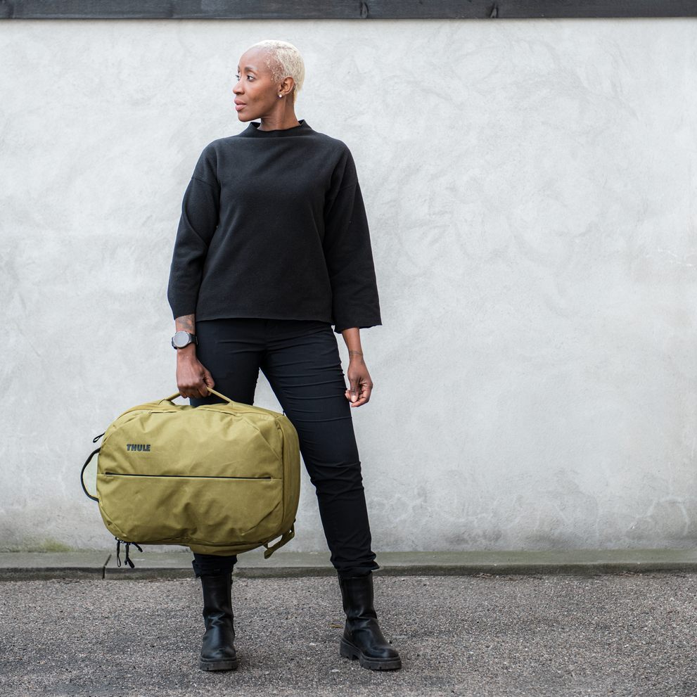 A woman with short blonde hair wearing black holds the tan Thule aion travel backpack.