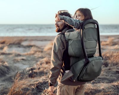 A father hikes with his daughter in a child carrier backpack on the beach.