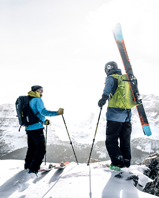 Two people stand on a mountain with skis and ski backpacks.